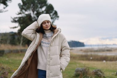 Açık hava ortamında kadın tarafından giyilen bej renkli kabarık ceket, örgü süveter ve şapka, rüzgarda savrulan saç, suyun yanında sakin bir alan, rahat stil havası, günlük kış moda sahnesi.