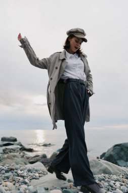 Geniş pantolonlu, beyaz gömlekli, gri ceketli ve kayalık bir sahilde yürüyen sıradan bir kadın. Bulutlu gökyüzü ve sakin okyanus arka planı, barışçıl ve şık açık hava havasını vurguluyor..