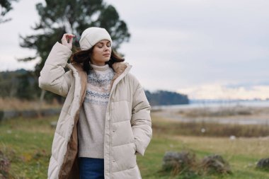 Sıcak giyimli kadın açık alanda poz veriyor, ağaçlar, serin tonlar ve sessiz gökyüzü, mevsimlik stil ve açık hava fotoğrafçılığı.