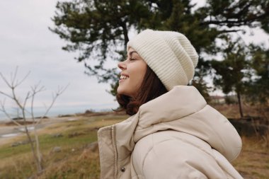 Sıcak bir palto ve bereyle gülümseyen bir kadının açık hava portresi, rüzgarlı bir manzarada doğayı kucaklayan deniz ve çam ağaçları, sakin ve huzurlu bir an.