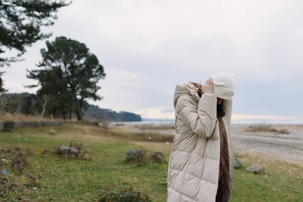 Açık hava sahnesinde, deniz kenarındaki çimenli bir uçurumun kenarında kabarık ceketli ve örgü şapkalı bir kadın, sakin bir ruh hali ve doğa, seyahat ya da yaşam tarzı fotoğrafçılığı için geniş bir gökyüzü yer alıyor..