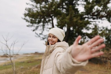 Samimi bir palto ve örgü şapkalı neşeli bir kadının yakın perspektif portresi, eller açık, geniş gülümseme, açık manzara, ağaçlar ve tarlalar yumuşak gün ışığında, davetkar, neşeli bir seyahat