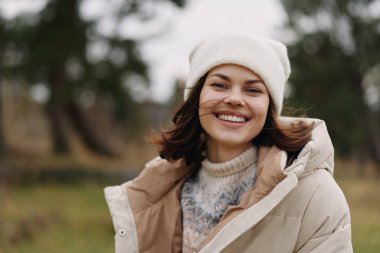 Açık kış ceketi ve örgü şapkası giyen gülümseyen bir kadının portresi, serin havalarda parkta dikilirken, neşeli bir stil ve açık hava sıcaklığı taşırken.
