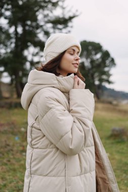 Açık havada kabarık ceketli ve rahat şapkalı bir kadının portresi, serin bir manzaranın ortasında sakin bir yüz ifadesi, sessiz tonlar ve rahat kış modası havası..