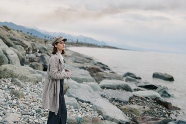 Paltolu ve şapkalı kadın sakin denizin kıyısında çakıl taşı sahilinde duruyor. Bulutlu gökyüzü ve uzak dağlar huzurlu bir atmosfer sağlar. Doğa kavramı, macera ve açık havada sessiz tefekkür..