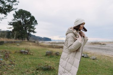 Kadın, palto, açık hava, rüzgar, manzara, moda, kış, manzara, sıcak bir kabarık ceketli yalnız bir gezgin esintili bir sahilde poz verir, seyrek ağaçlar, yumuşak bulutlar, sakin bir ruh hali, kentsel olmayan moda anı