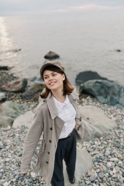 Paltolu ve şapkalı gülümseyen genç kadın sakin deniz kenarında çakıl taşı kumsalında poz veriyor. Rahatlamış sonbahar stili, doğa arka planı ve doğal ışıkla çekilmiş neşeli açık hava yaşam tarzı..