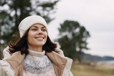 Açık hava portresi, kışın giyinmiş gülümseyen bir kadın bej ceket ve beyaz örgü şapkalı rahat bir süveter, rüzgarlı bir tarlaya ve uzak bir ufka karşı.