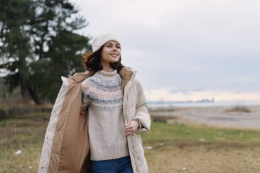 Açık hava portresi, deniz kenarındaki sıcak süveter ve ceketli, rahat bir hava için çimen, ufuk ve kıyı manzaralı gülümseyen bir kadın portresi..