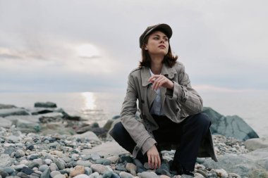 Gün batımında çakıl taşı sahilinde çömelmiş şapka giyen sıradan bir genç kadın. Bulutlu gökyüzü ve kayalar yansıma, sıradan moda ve doğal manzara için sakin bir atmosfer yaratıyor..