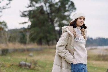 Açık hava moda sahnesi, yastıklı bir kadın, örgü süveter ve bere, rahat poz açık çayırlar ve uzak ağaçlar, rahat mevsim tarzı