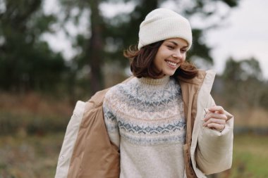 Açık hava portresi, rahat bir süveter ve bere giymiş bir kadın, bir ceketin üzerinde kayarken gülümsüyor, doğal manzara ve nazik, bulutlu gökyüzü ile çevrili..