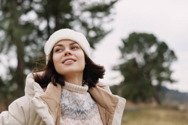 Açık havada bir kadın portresi, gülümseyen, bereli ve kalın bir palto giyen, arka planda ağaçlar, kışın serin atmosfer, sakin ifade, günlük moda ve doğal güneş ışığı..