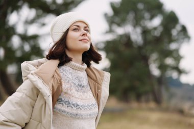 Sıcak örülü bir süveter ve açık havada rahat bir palto giyen bir kadının portresi, gözler kaldırılmış, yumuşak manzaralı ve arka planda uzak ağaçlarla dolu, narin bir ışıkla yıkanmış, gevrek bir günün tadını çıkaran bir kadın..