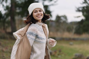 Örgü ören bir süveter ve palto giyen gülen bir kadın, açık hava sonbahar ortamında bereli, elinde açık renk bir ceket, hafif bir güneş ışığı, gündelik moda ve rahat ruh halini ön plana çıkarıyor..
