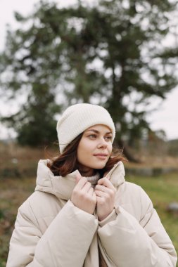 Kabarık bej ceket ve açık bir manzarada beyaz örgü şapka giyen bir kadının portresi, sakin bir ruh hali, hafif bir esinti, rahat kış havası ve açık hava yaşam tarzı.