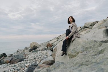 Siyah saçlı sıradan bir kadın bulutlu gökyüzünün altında sakin suların yanında kayalık bir kıyıda oturuyor. Sahil manzarasında huzur, yansıma ve doğal çevre kavramı.