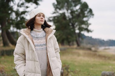 Kışın giyinen kadın, açık hava sahnesi, yumuşak ışık, bej ceket ve örgü süveter, bere, tarla ve ağaçlar, yalnızlığı yakalayan sakin bir portre ve doğa ile sessiz bir bağ.