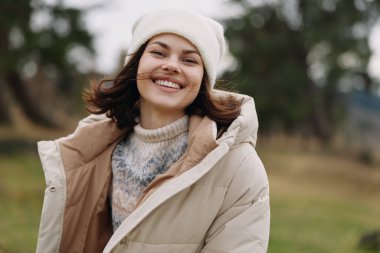 Sıcak bej ceket giyen ve açık havada kazak ören gülümseyen bir kadın, serin havada bir park ortamının keyfini çıkaran, günlük tarzını ve iyimser tavırlarını hayat tarzı fotoğrafında vurgulayan bir kadın..