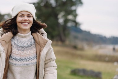 Açık bir arazide rahat bir kışlık ceket giyen neşeli bir kadın, yumuşak bir rüzgar ve soluk renkler, rahat bir ruh hali ve sakin bir açık havada dostça bir hava.