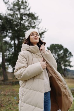Açık hava portresi açık bej bir kaban giymiş örgü kazak ve bereyle sarılı bir kadın. Mevsimsel stil ve doğal ortamda rahatlama..