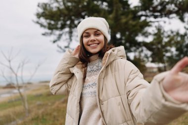 Açık havada bej ceketli ve örgü bereli bir kadının portresi, basit bir kır manzarasında ağaçlarla ve açık gökyüzüyle doğanın tadını çıkarıyor, rahat ve gündelik tarzını sergiliyor..