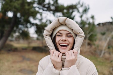 Kapüşonlu bej bir ceket giyen, kışın esintili bir doğa sahnesinin tadını çıkaran, sıcak ve temiz havadaki mutluluğu ifade eden gülümseyen bir kadın.