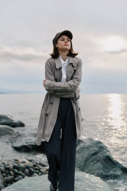 Şık bir palto ve şapka giyen şık bir kadın sakin denizin yanındaki kayalık sahilde poz veriyor. Akşam ışığı ve bulutlu gökyüzü açık hava tarzını ve barışçıl doğa düzenini vurguluyor.
