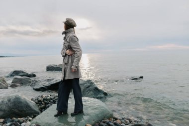 Gün batımında bulutlu gökyüzü ile birlikte sakin okyanusun kıyısındaki taşların üzerinde tek başına duran palto ve şapka. Yalnızlık kavramı, doğa, barışçıl yansıma, ve açık havada su keşfi..