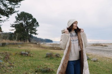 Dışarıdaki kadın, kalın bir palto giyiyor ve kazak örüyor. Su kenarında şapka takıyor ve çimen takıyor. Duruşu gün batımında sakin bir kırsalda dinleniyordu.