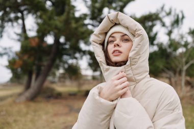 Kapüşonlu kışlık bir ceketin, sakin bir kadın modelinin üzerinde, açık havada parkta, yumuşak doğal ışık dokusu ve rahat bir ruh hali..