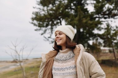 Kış kıyafetleri içinde gülümseyen kadın ağaçların yanında duruyor ve sakin bir kıyı şeridi, bereli ve açık hava sahnesinde açık bir ceketin altına örgü örüyor..