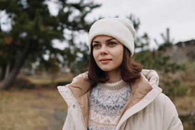Açık hava sahnesi, açık renk ceketli ve örgü şapkalı sakin bir kadın, doğa ile çevrili serin bir tonu ve sakin bir havası var..