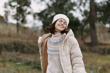 Açık hava sahnesinde kabarık montlu ve örgü kazaklı neşeli bir kadın, bereli, serin havada parkta duruyor, rahat bir stil ve parlak bir ifade sergiliyor..