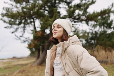 Sade bej ceketli ve örgü şapkalı genç bir kadının açık hava portresi. Çıplak ağaçlar ve yumuşak ışıklarla dolu bir parkta temiz havanın tadını çıkarıyor. Huzur ve keşif yapıyor..