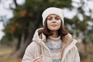 Kışın bej ceketli ve örgü süveterli bir kadının portresi, beyaz bir bere takıyor, açık havada yumuşak ağaçların arasında, sıcaklık yayıyor, sade bir tarz ve rahat bir ruh hali içinde.