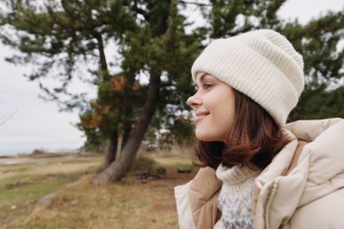 Açık havada bej ceketli ve yumuşak örgü şapkalı bir kadının portresi, ağaçlar ve çimenlerle dolu sakin bir kırsal bölgeye bakıyor, doğal hava koşullarında huzurlu ve düşünceli bir ruh hali sergiliyor..