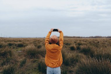 Kafasının üstünde akıllı bir telefon tutan yaşam tarzı kadın doğal bir çim tarlasında manzara yakalıyor. Gündelik giysiler ve sonbahar renkleri açık hava macerası ve özgürlüğü vurguluyor.