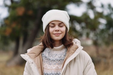 Kış modasında rahat bir kadın portresi, katmanlı örgü kazak ve yastıklı ceket, huzurlu bir park ortamında beyaz bere ve huzurlu bir ifade, rahatlık ve stil taşımaktadır..