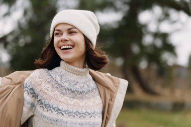 Açık havada örgü süveter ve bereli gülümseyen bir kadının portresi, açık renk ceketli, serin bir sonbahar gününde, hafif bir esintiyle bir parkta.