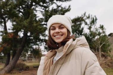 Açık havada, kabarık ceketli ve bereli neşeli bir kadının portresi, çıplak ağaçlarla park yeri, rahat bir mevsim tarzı ve samimi bir yaşam tarzı..