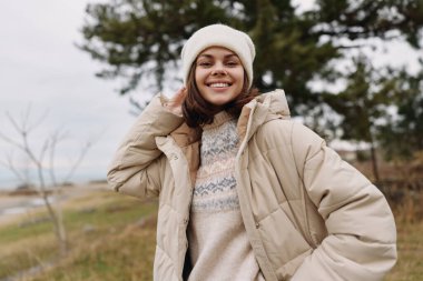 Açık bir tarlada duran, rahat bej bir ceketle gülümseyen kadın, örgü ören kazak ve şapka, rahat açık hava modası, doğa havası ve rahat kış fotoğrafçılığı tarzı..
