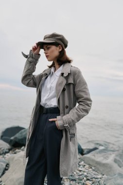 Dokulu palto ve eşleşen şapka giyen genç bir kadın okyanus yakınlarındaki kayalık kıyılarda düşünceli bir şekilde poz veriyor. Sonbahar kıyafetleri, gündelik tarz ve karamsar doğal ışıklandırma içeren moda konsepti.