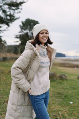 Açık hava sahnesinde kıyı şeridi yakınlarındaki bir parkta ceketli, kazaklı, bereli ve sıradan görünümlü gülümseyen bir kadın var. Doğal ışık, sakin ve neşeli yaşam tarzı