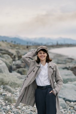 Kareli bir palto ve şapka giyen mutlu kadın, dağların ardında bulutlu gökyüzü olan kayalık bir sahilde poz veriyor. Rahatlamış sonbahar stili ve doğa keşfi havası açık havada yakalanmış..