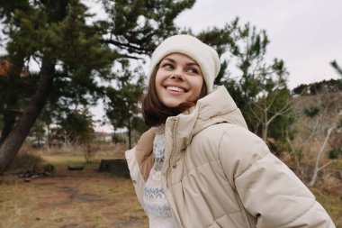 Yumuşak bej ceketli ve örgü şapkalı neşeli bir kadının portresi, çıplak ağaçlarla dolu bir parkta duruyor, sıcaklığı, rahatlığı ve mevsimsel havasını iletiyor.