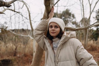 Açık havada, çıplak bir ormanda bir ağaca yaslanmış kalın bir palto ve bere giyen sakin bir ruh hali, doğal dokular ve soğuk havada sakin bir tefekkür hissi taşıyan bir kadının portresi..