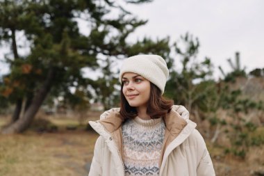 Açık havada, rahat bir palto ve örgü süveter giyen bir kadının portresi, yumuşak kış ışığı altında bir parkta duruyor, rahat bir hava ve rahat bir hava sergiliyor..