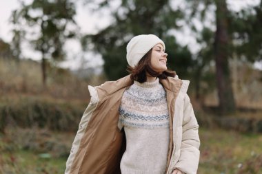 Açık bir parkta bej ceketli bir kadın ve örgü örüyor. Ağaçlı bir parkta. Yüz ifadesi rahat ve memnun..