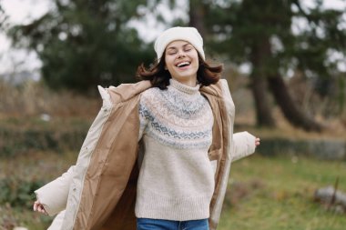 Açık hava sahnesinde günlük tarz ve mutluluk, rahat bir ceket ve örgü süveter içinde gülümseyen bir kadın, temiz hava, hareket ve kaygısız ruh halini kucaklıyor.